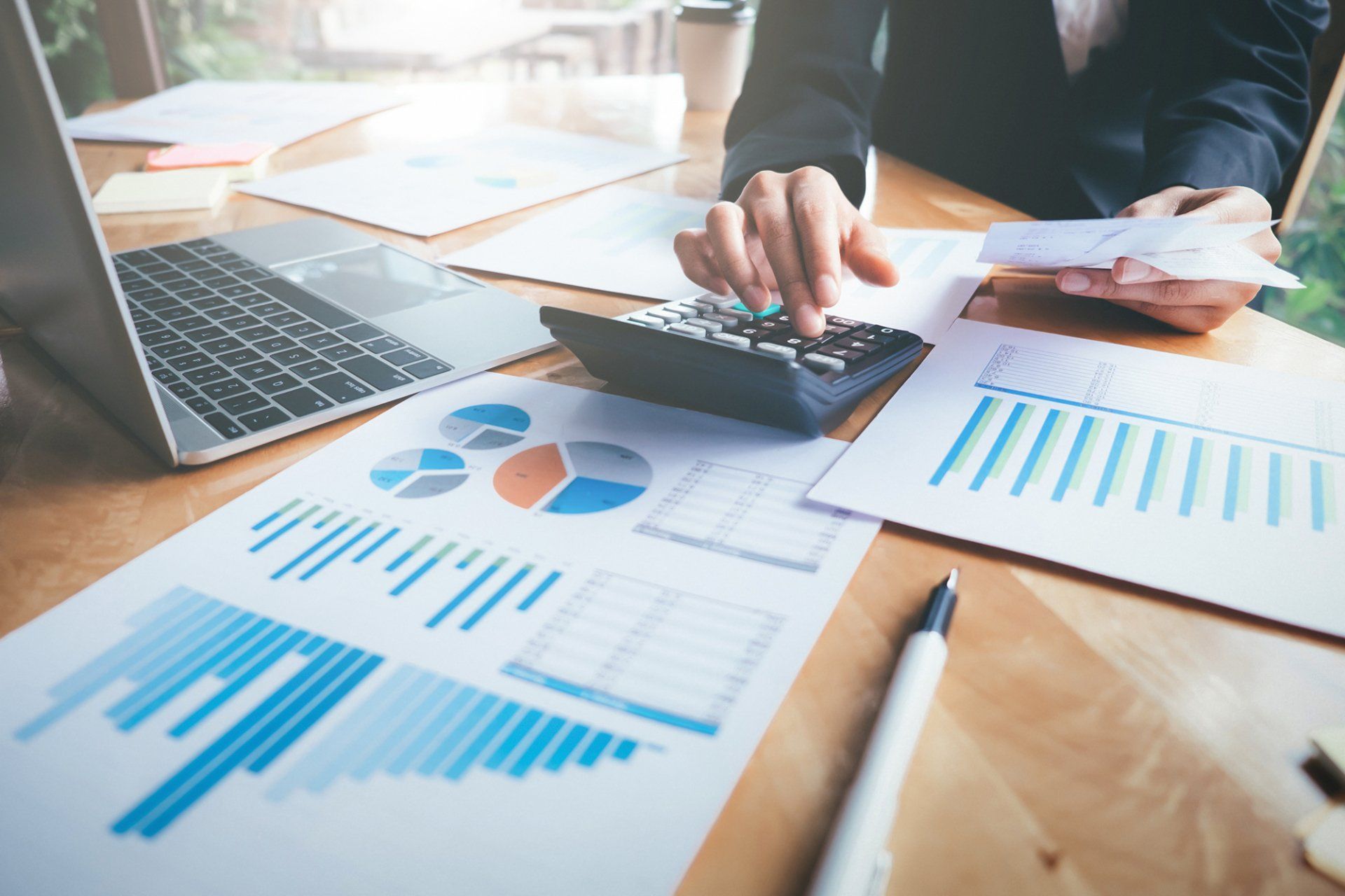 Person using a calculator at a desk with financial charts, a laptop, and documents.