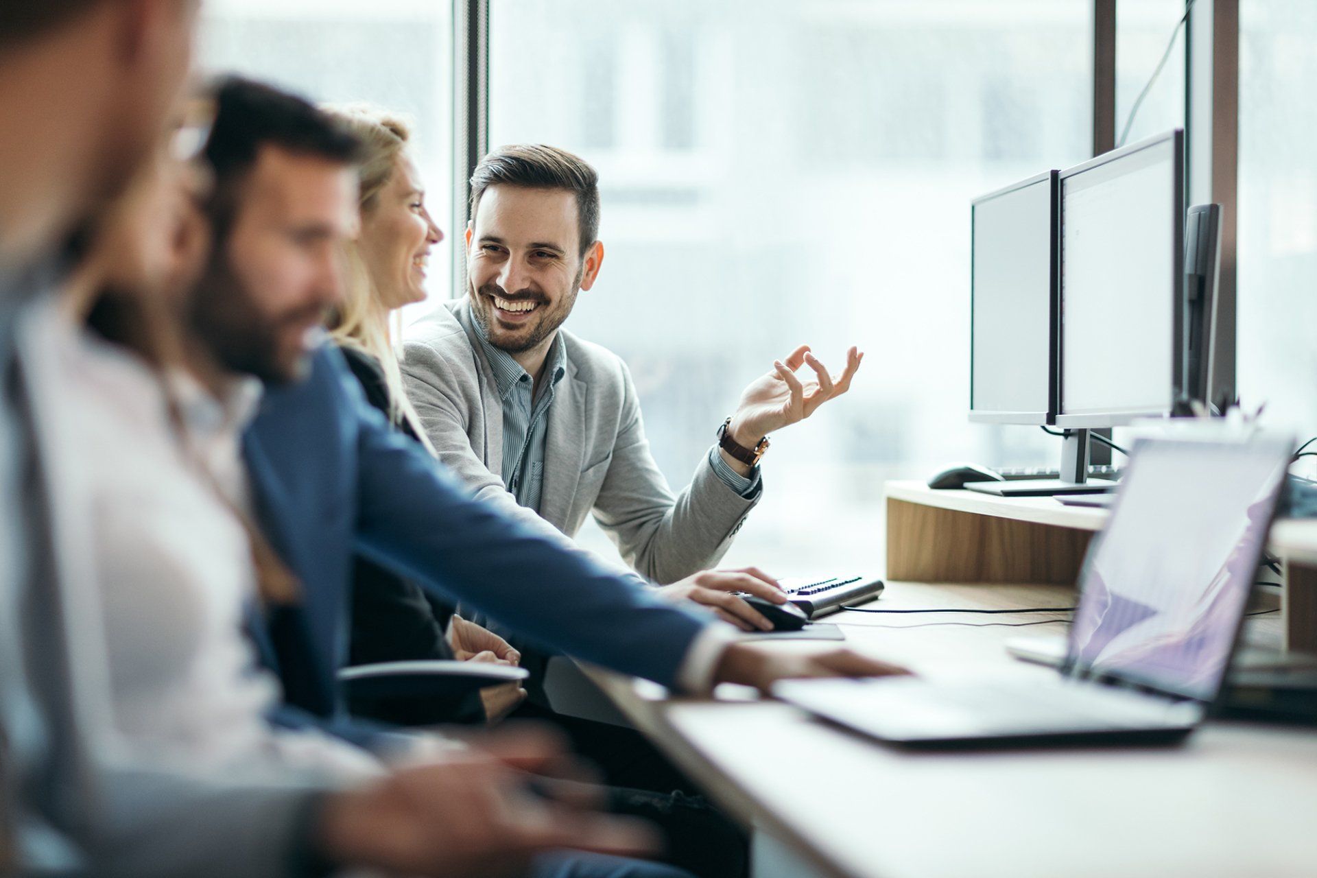 Four people collaborating at a desk in an office. Man smiles, gestures with his hand. Windows in background.