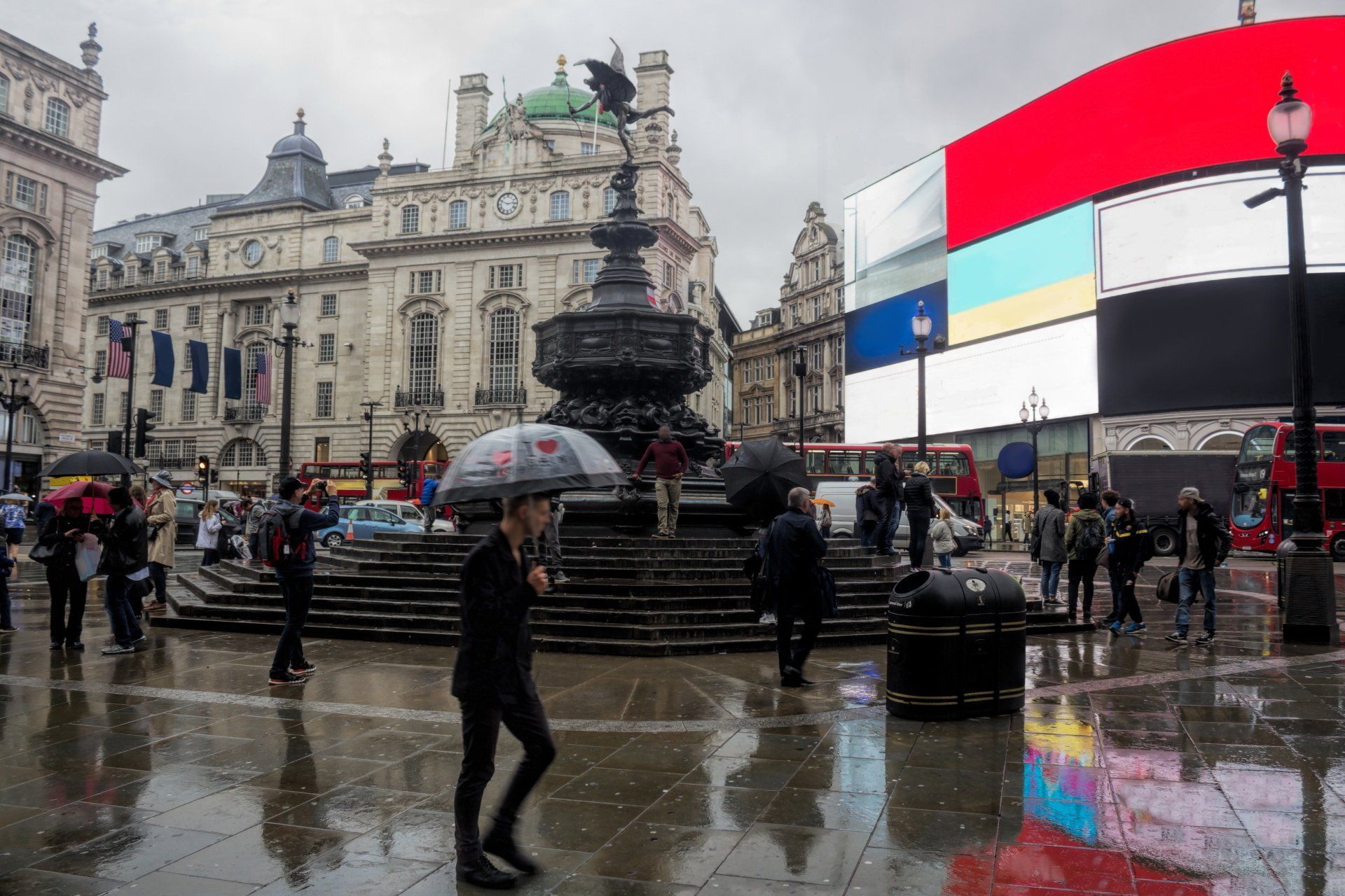 Piccadilly Circus, London, rainy day; crowds around Eros statue, illuminated billboards.