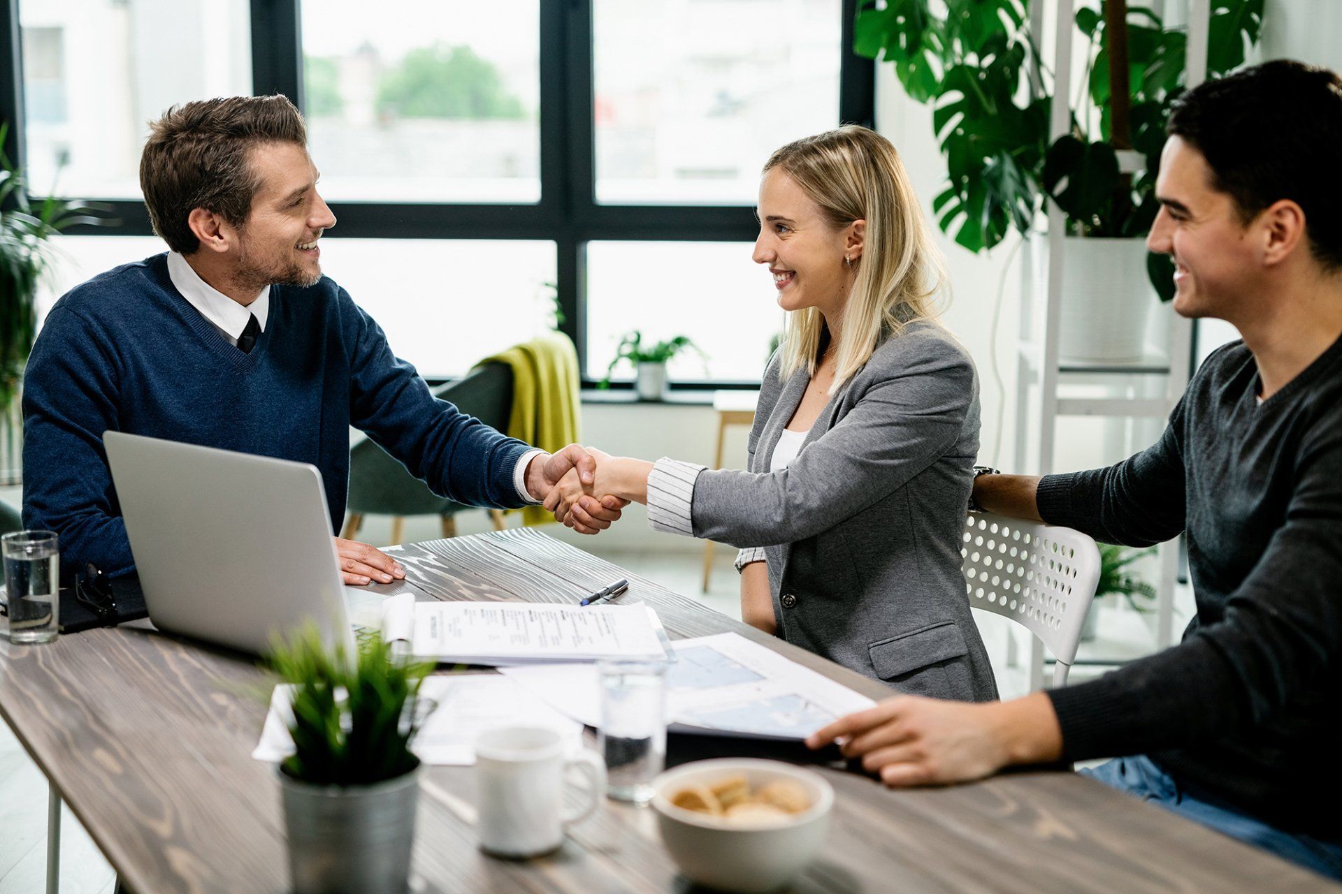 Three people, two women and a man, shake hands at a table in an office setting.