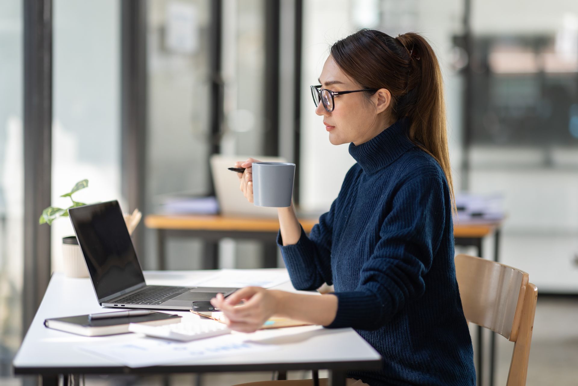 Woman with glasses in blue sweater working on a laptop, holding a mug in an office.
