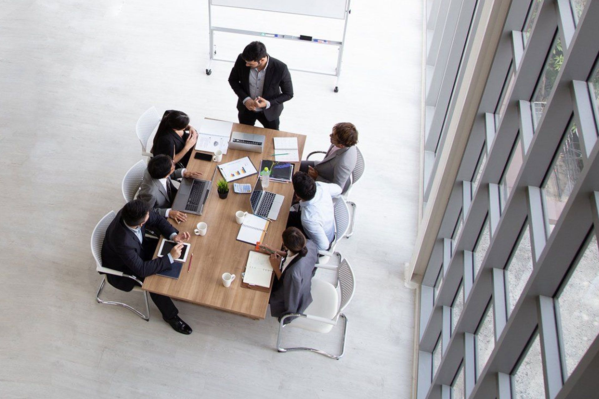 Business meeting with diverse people seated at a table; one man standing, presenting. Bright room with large windows.
