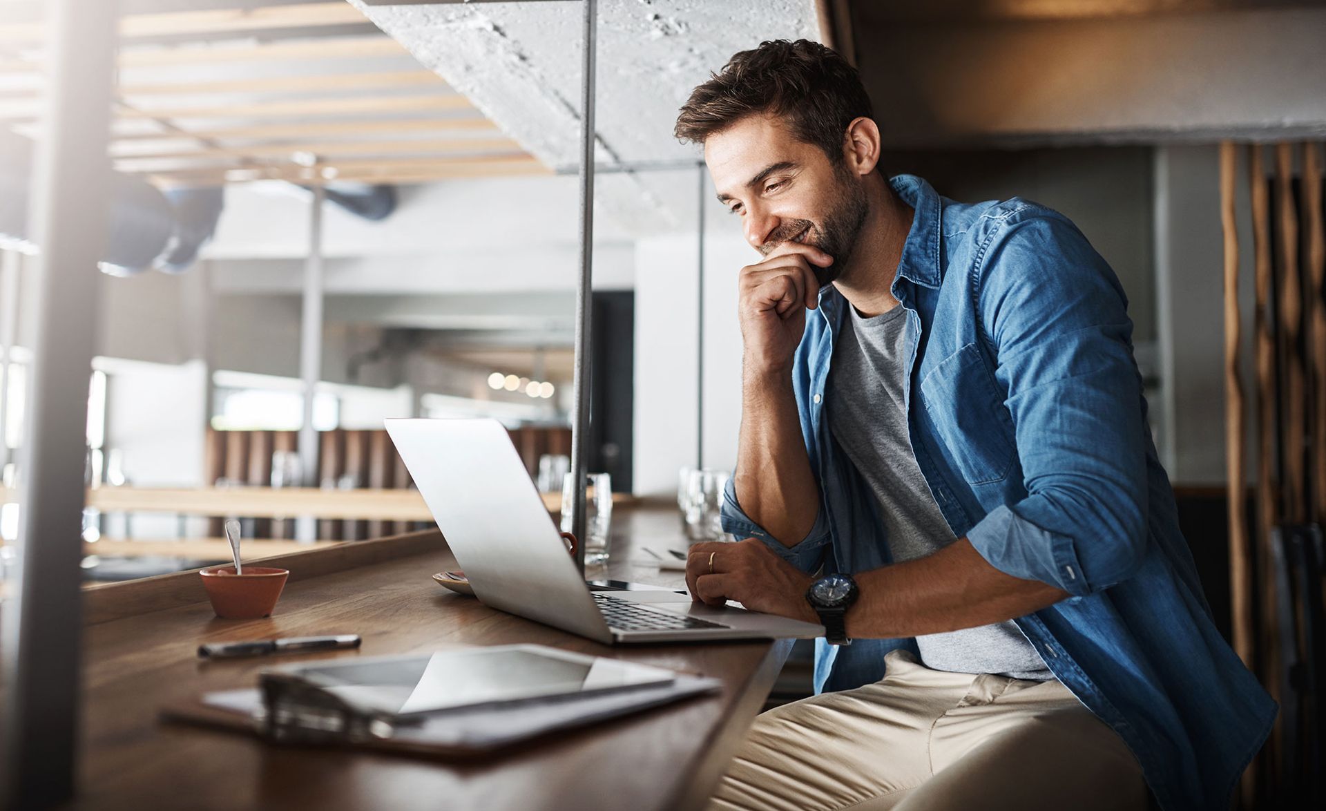 Man working on laptop at a counter, looking thoughtful, wearing a denim shirt.