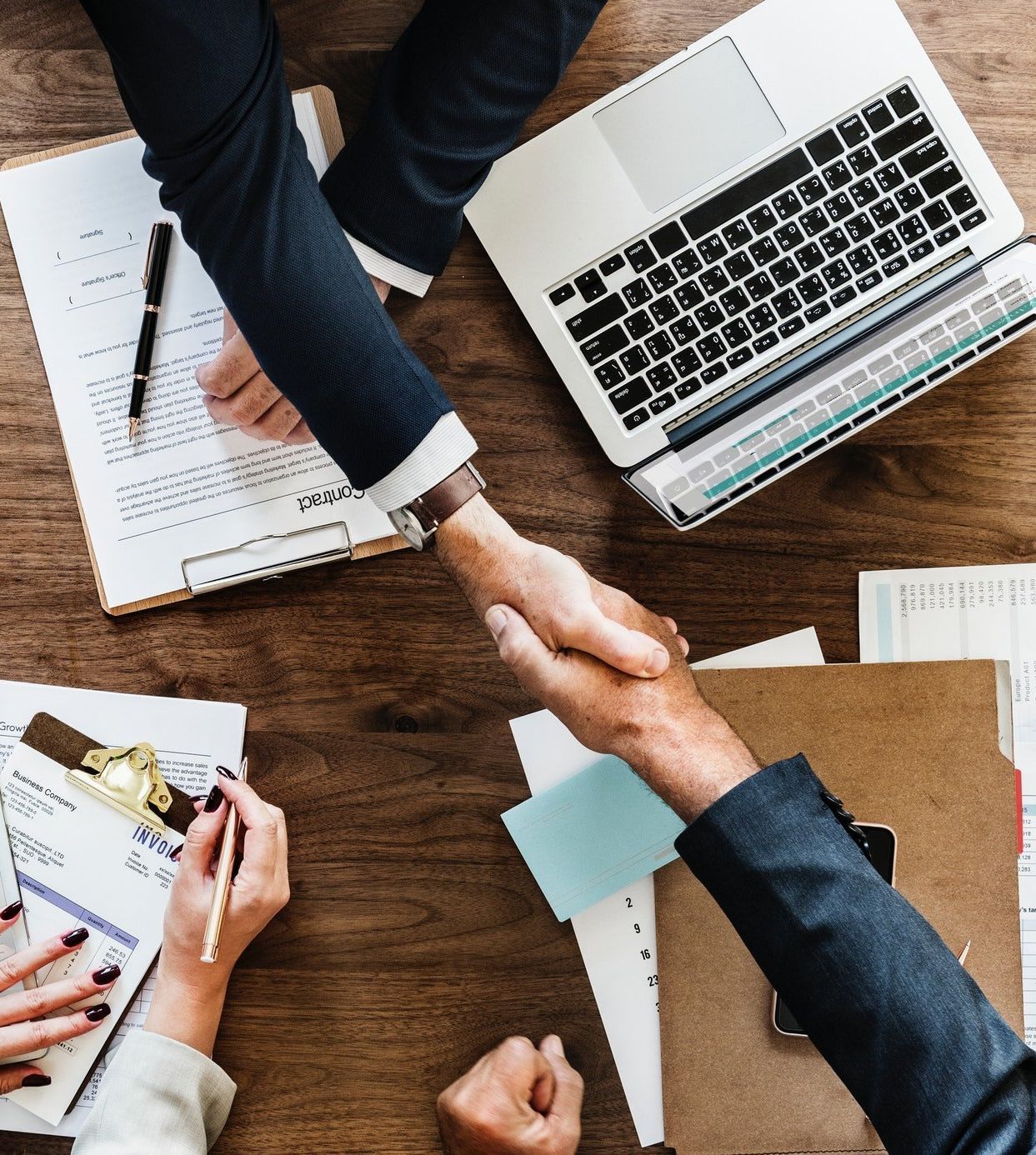 Two people in suits shake hands over a wooden table with documents and a laptop.