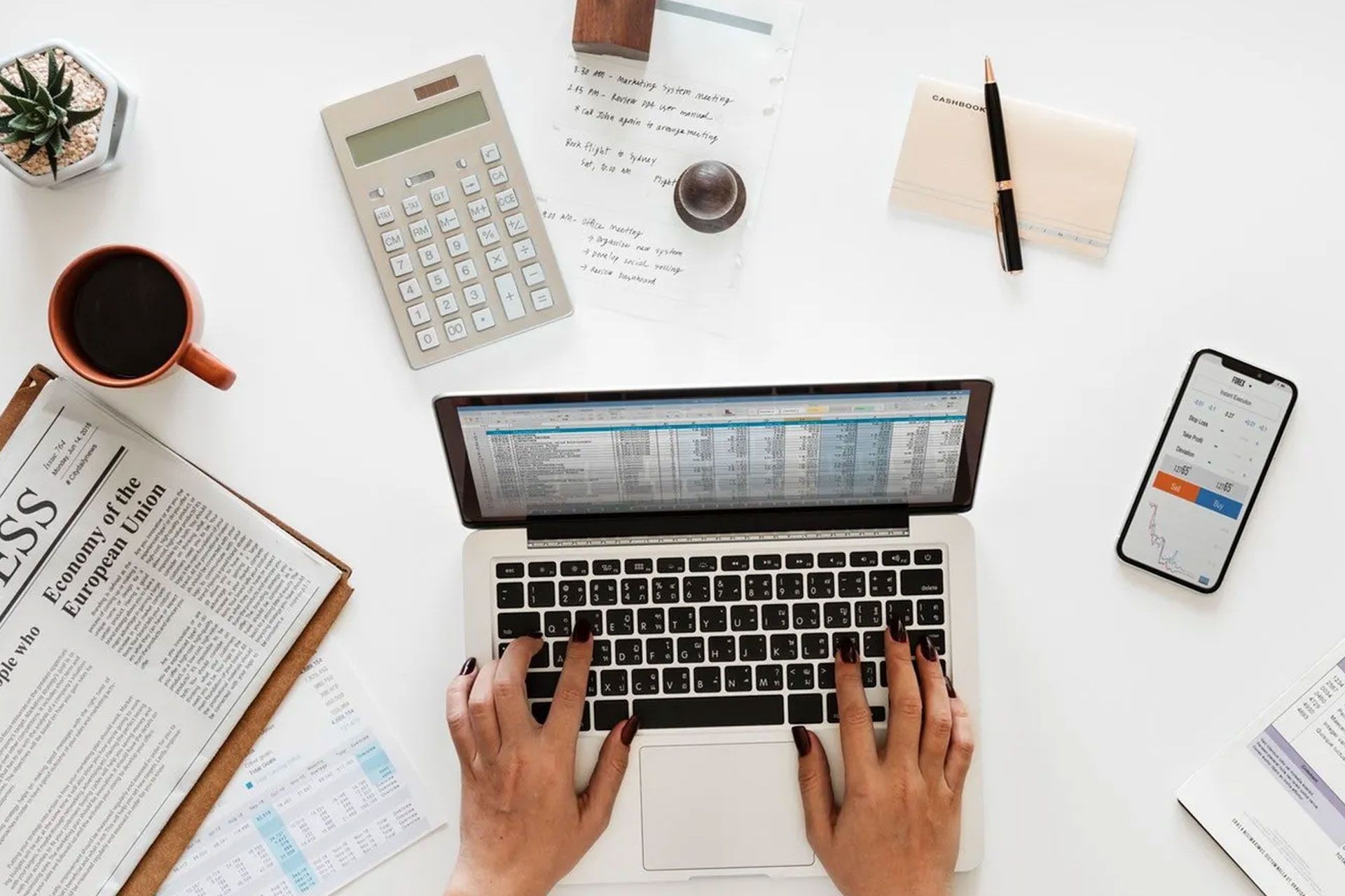 Person typing on laptop with spreadsheet; desk with calculator, phone, coffee, newspaper, and notepad.