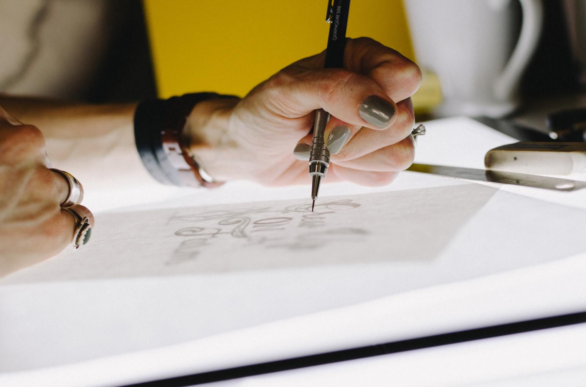 Hand with pencil drawing on a lit-up drawing pad. Grey nails, rings, and a watch visible.