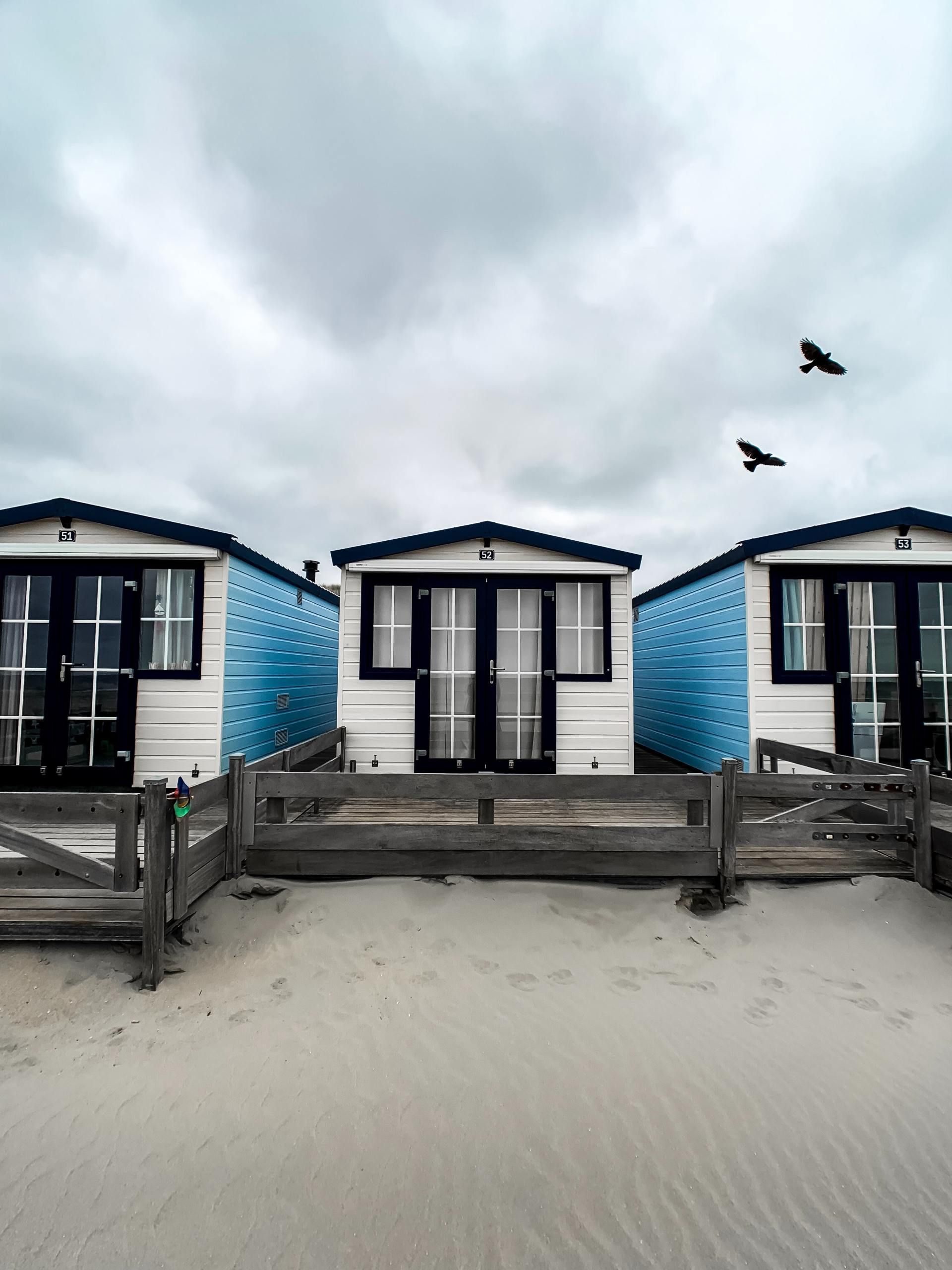 Three colorful beach huts on a sandy beach under a cloudy sky, with two birds flying above.