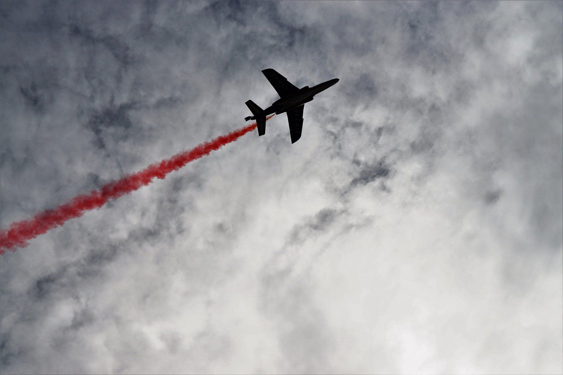 Jet airplane silhouetted against cloudy sky, trailing red smoke.