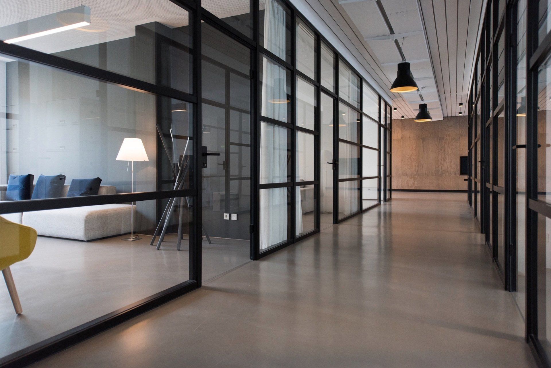 Modern office hallway with glass-walled rooms, concrete floor, and black-framed doors. Includes couch, chairs, and lights.