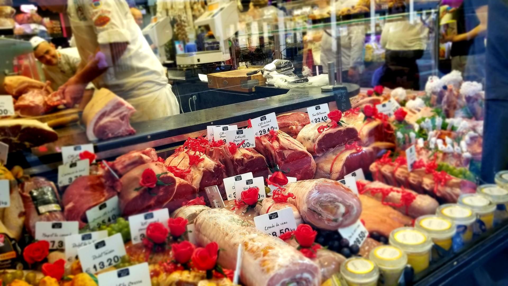 Butcher shop display case filled with various cuts of meat, red, pink, and white. Butcher in white apron preparing meat.