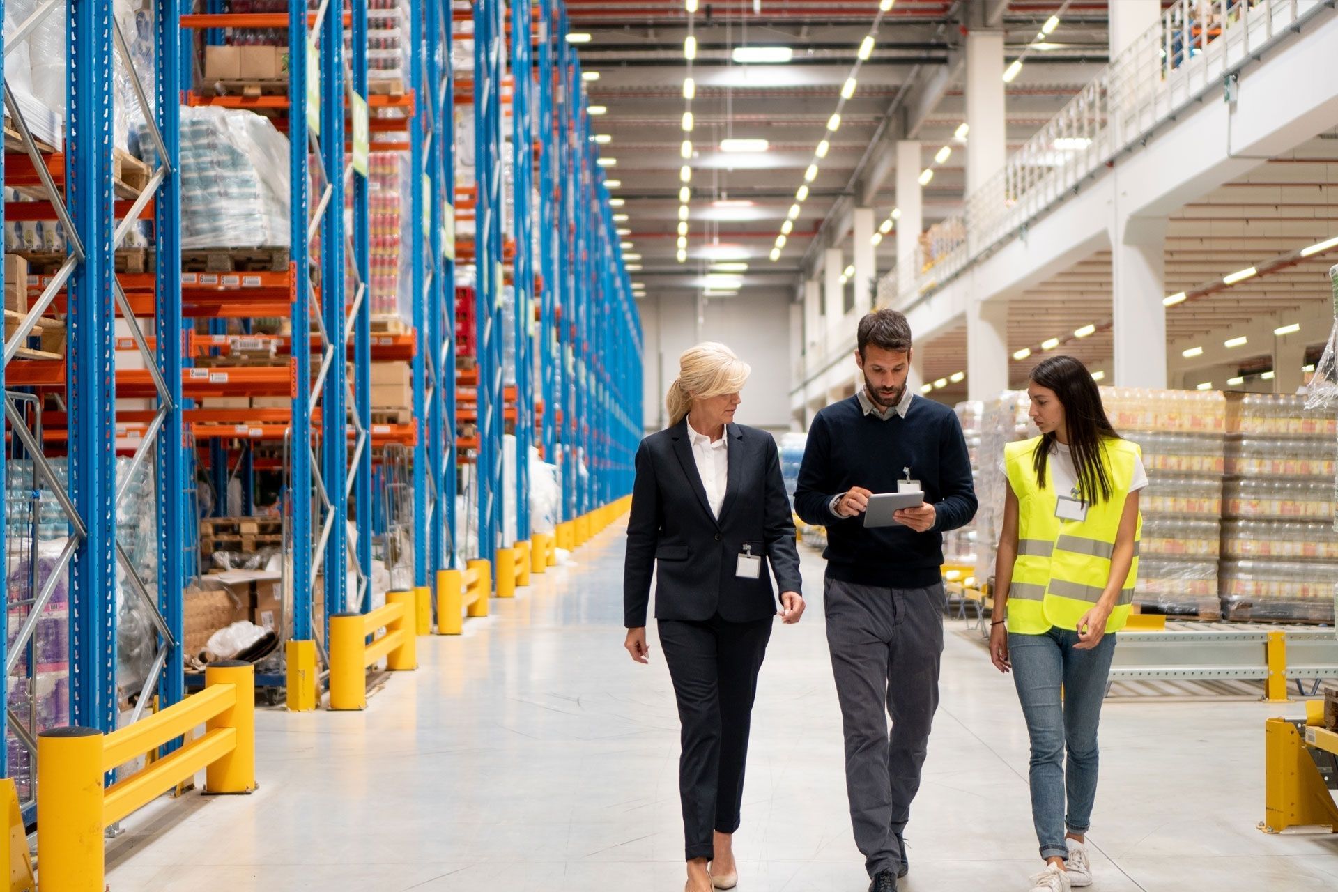 Three people in a warehouse: a woman in a suit, a man with a tablet, and a woman in a safety vest walk down an aisle.