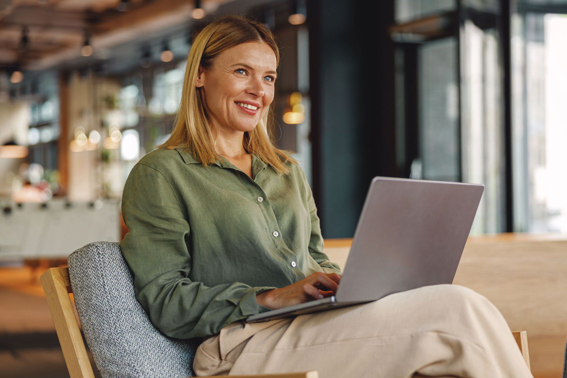 Woman with blonde hair smiles while working on a laptop in a well-lit modern space.