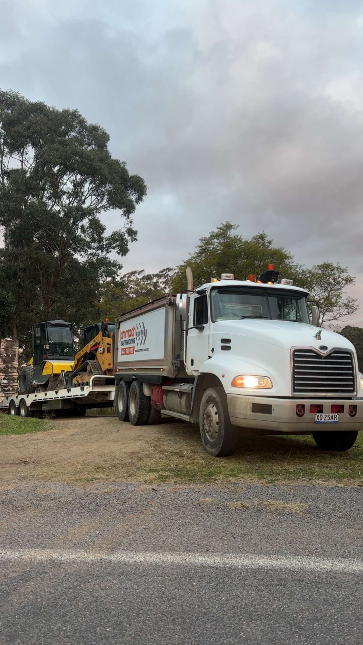 White truck with trailer transporting yellow compact loader and roller, parked on a rural roadside — Jammach Earthmoving in Nulkaba, NSW