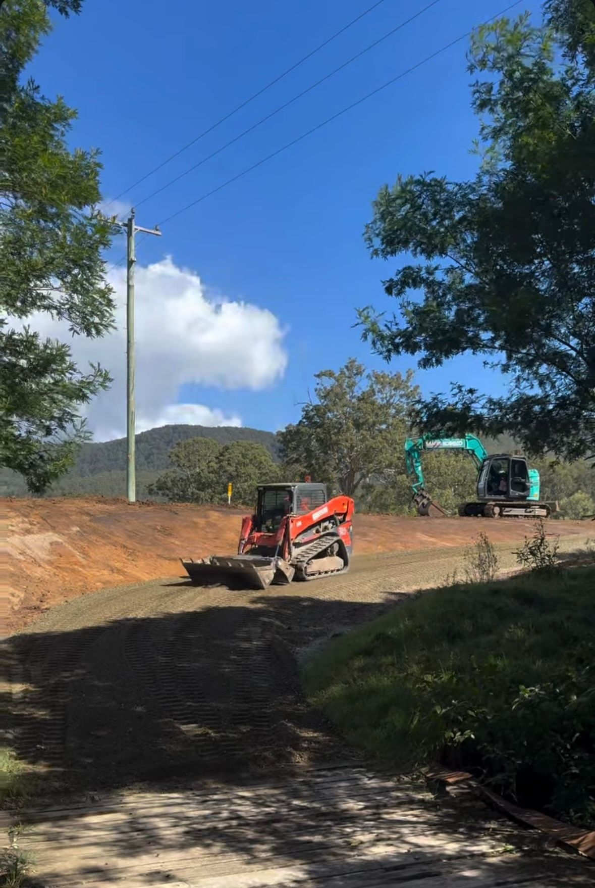 Red skid steer loader and teal excavator working on a dirt road in a rural area with trees and hills in the background. — Jammach Earthmoving in Nulkaba, NSW