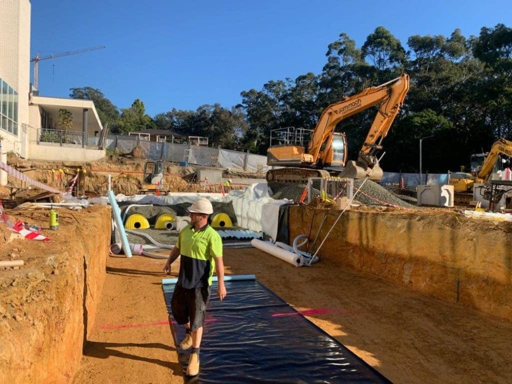 A Man is Walking Through a Construction Site With a Large Excavator in the Background — Jammach Earthmoving in Newcastle, NSW