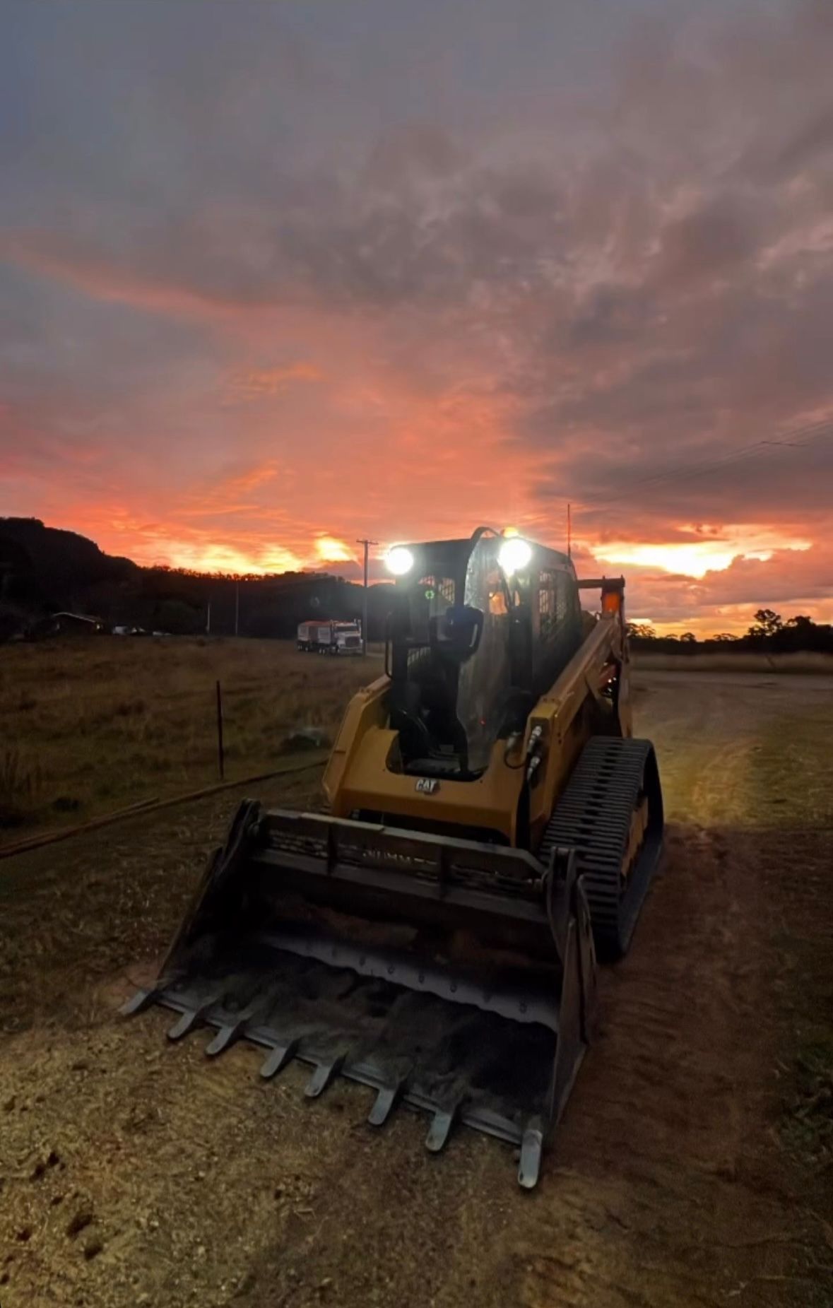 Yellow compact track loader with headlights on, parked on dirt at sunset with dramatic sky — Jammach Earthmoving in Nulkaba, NSW