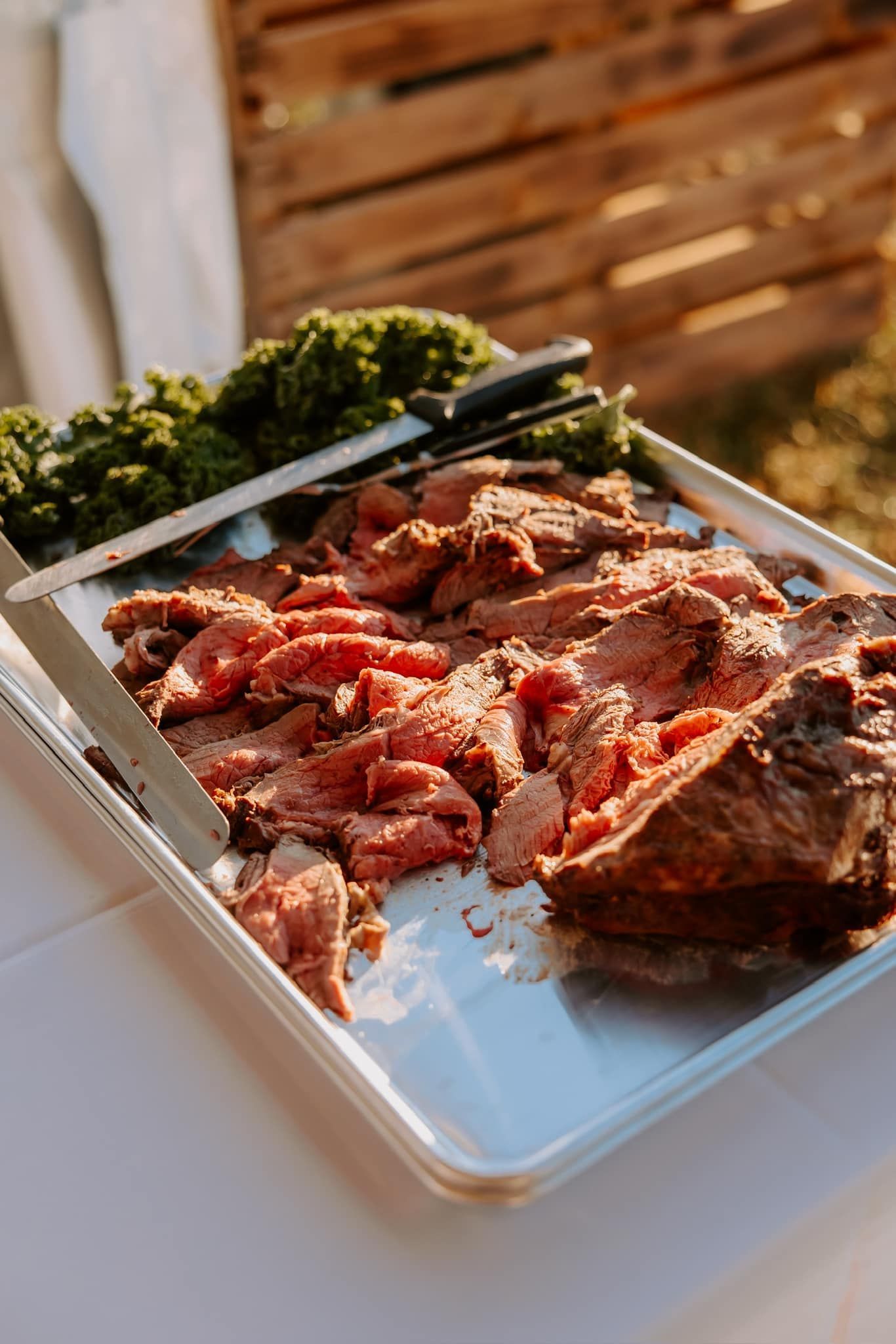 Sliced roast beef on a metal tray, garnished with green garnish and serving utensils.