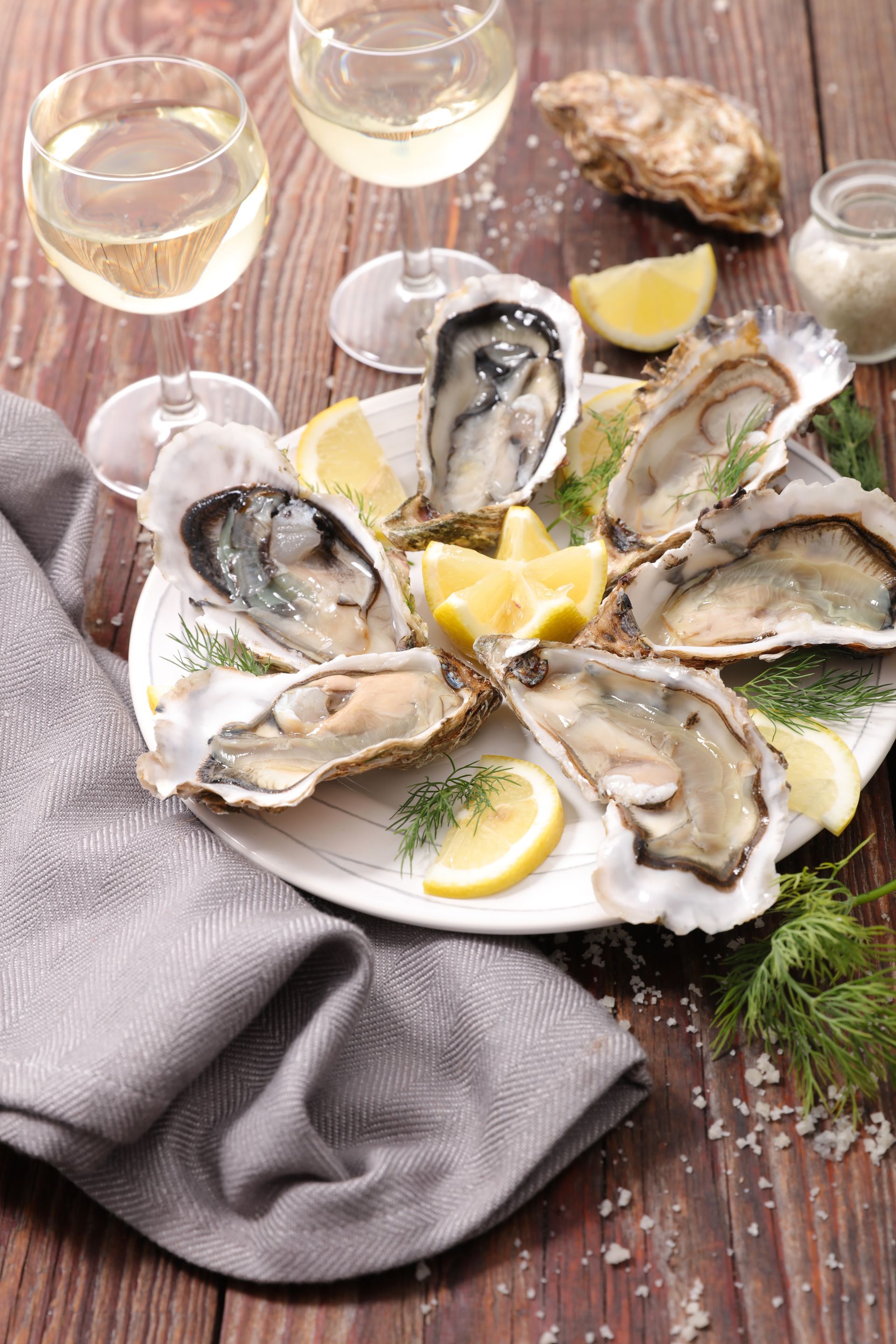 Plate of raw oysters with lemon slices, dill, and white wine glasses on a wooden table.
