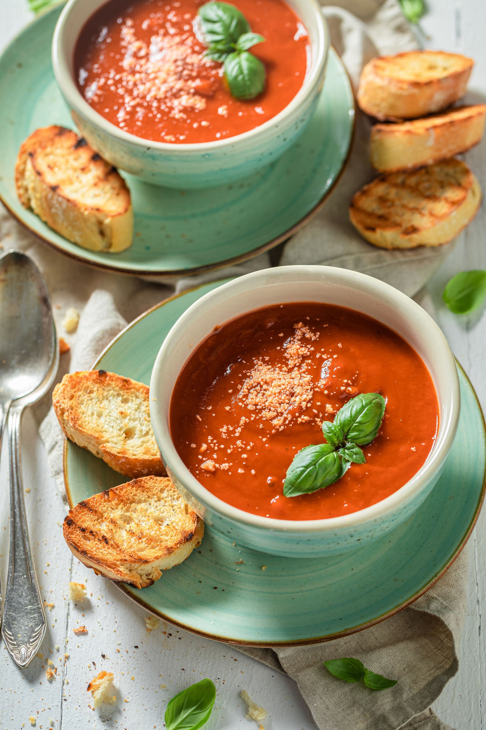 Two bowls of tomato soup with grilled bread, garnished with basil and parmesan.