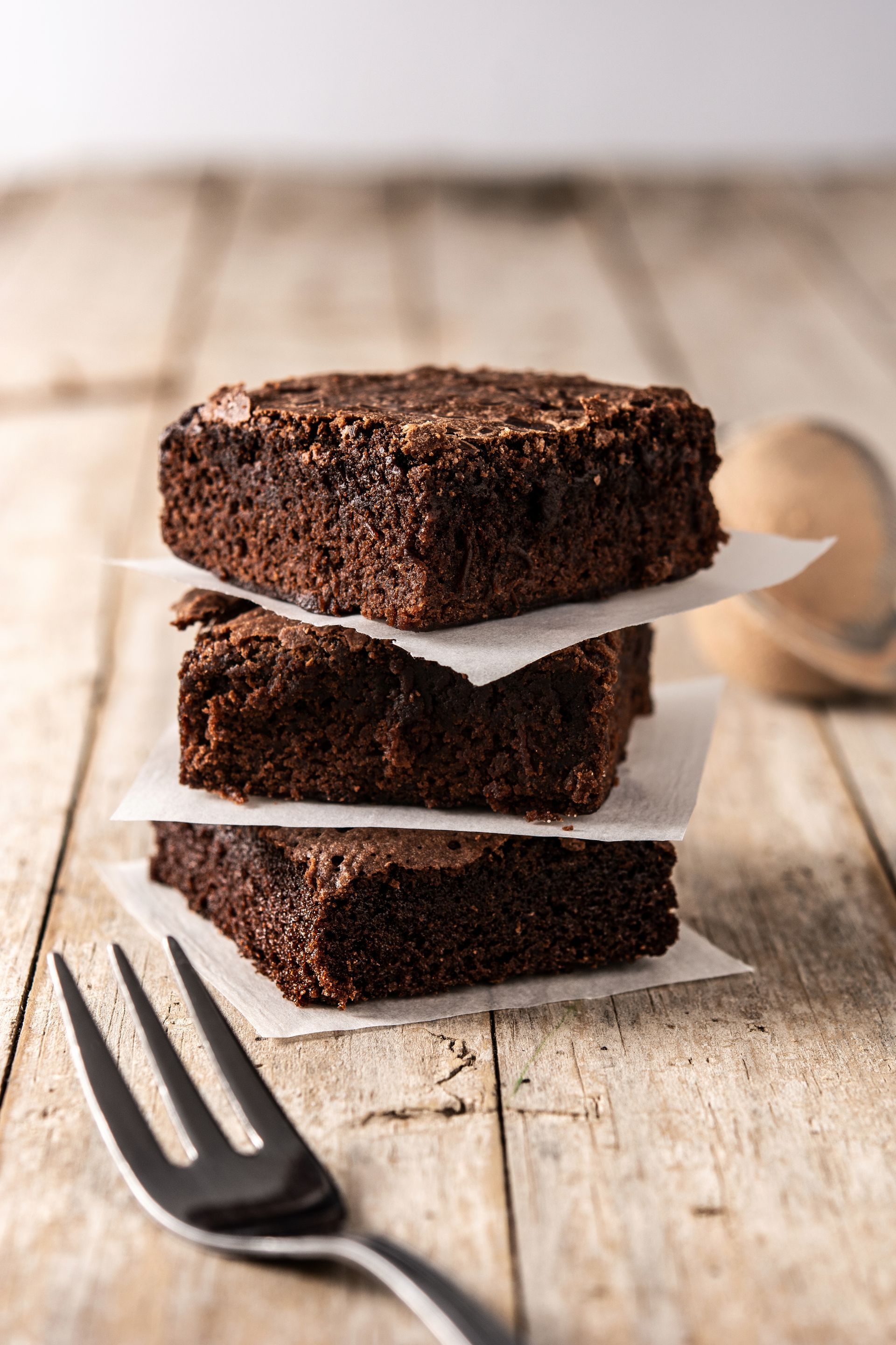 Stack of three dark chocolate brownies on parchment paper, next to a fork on a wooden table.