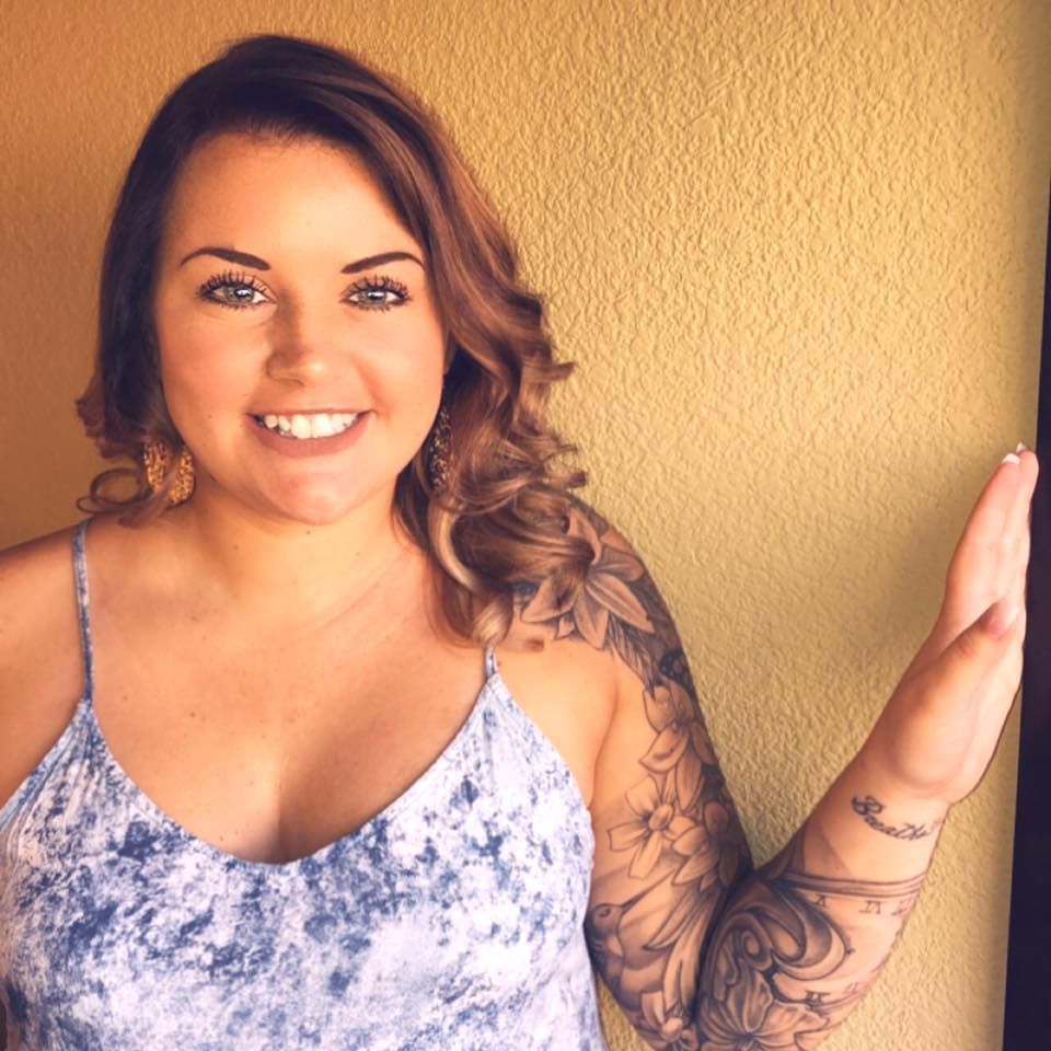 Woman with curly brown hair, floral tattoo, smiling, in blue top, hand up by yellow wall.