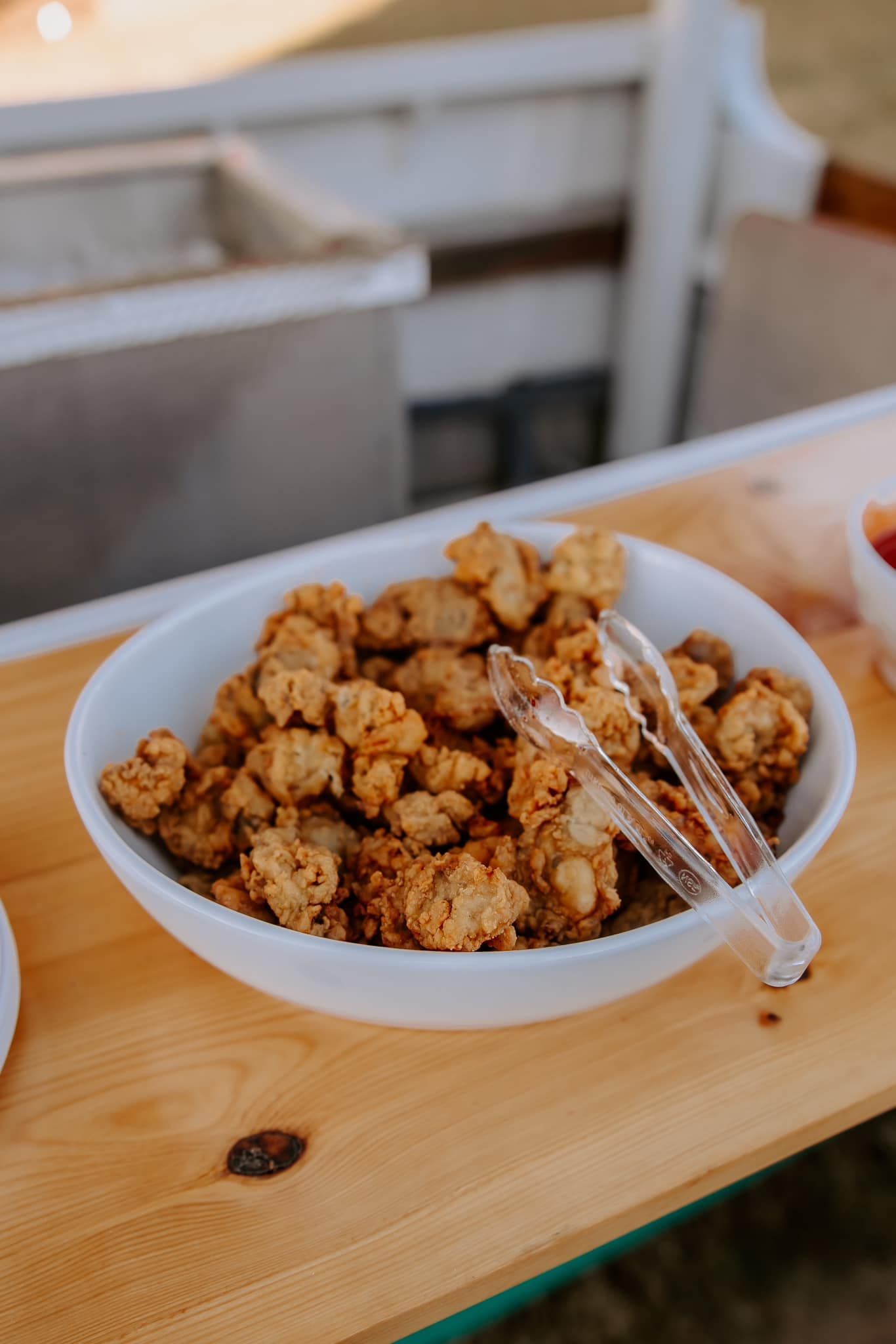 Fried food in a white bowl with serving tongs on a wooden table, outdoors.
