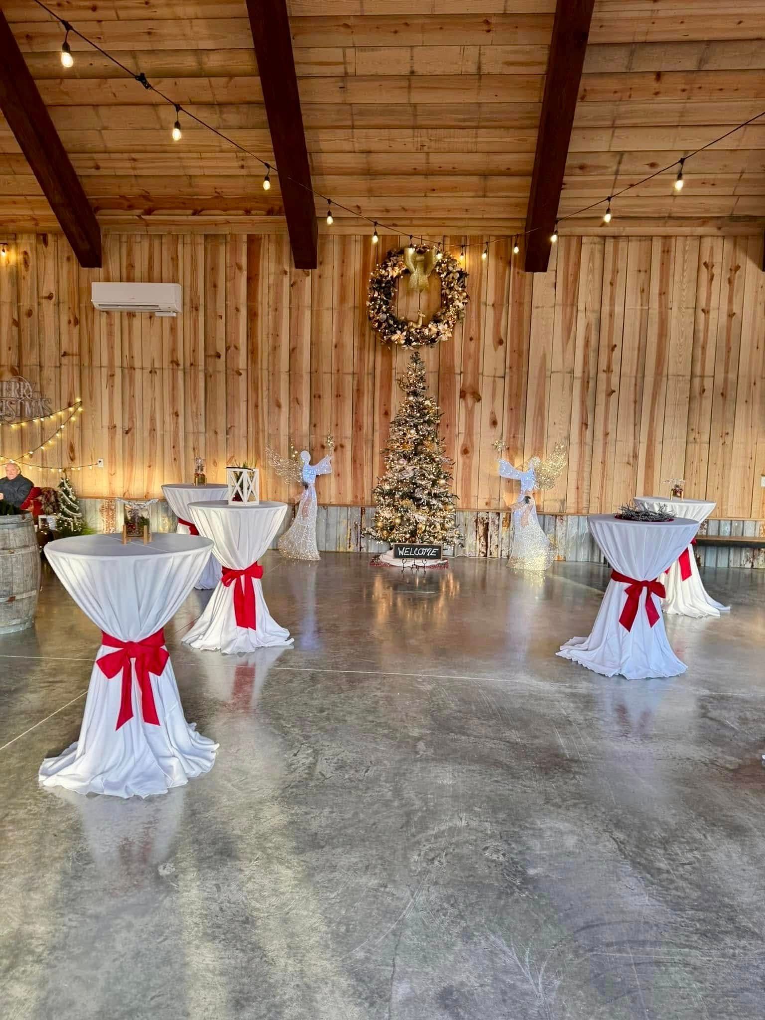 Festive indoor event space with cocktail tables, Christmas tree, and wreath; wooden walls and gray floor.