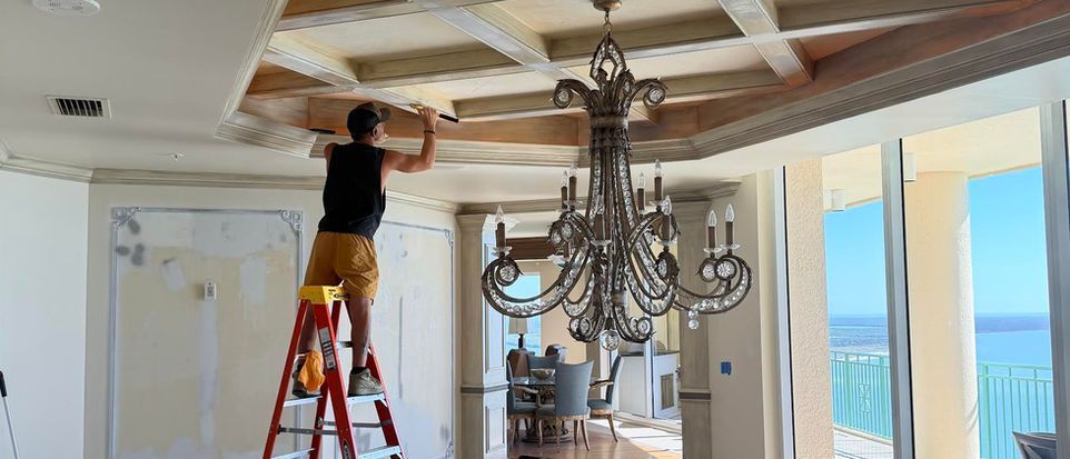 Person on a ladder installing crown molding in a bright room with a chandelier and ocean view