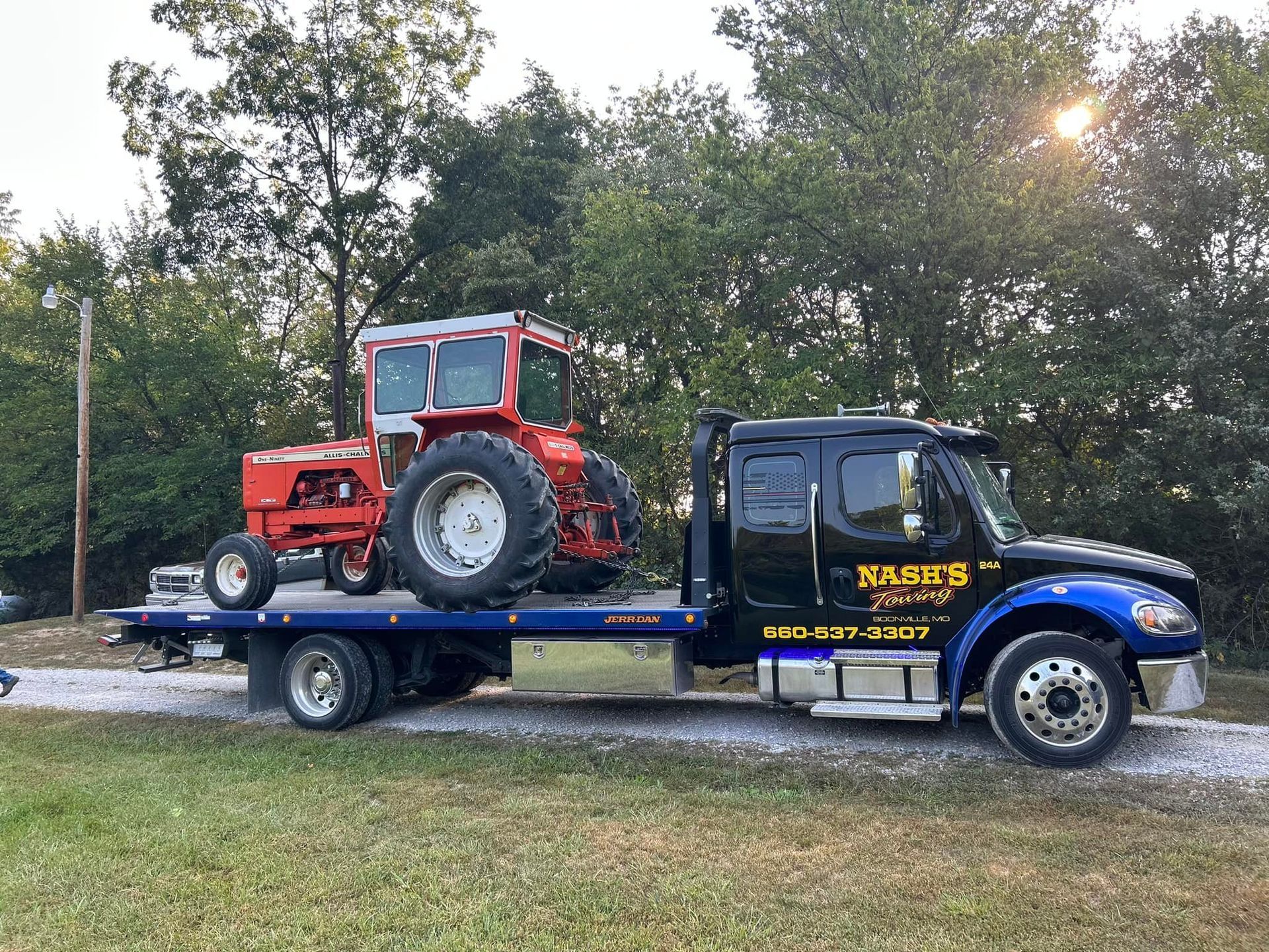Red tractor on a flatbed tow truck; parked on a gravel road, with trees in the background.
