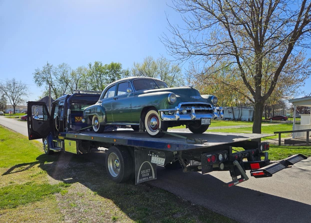 Green classic car on a tow truck, parked outdoors on a sunny day.
