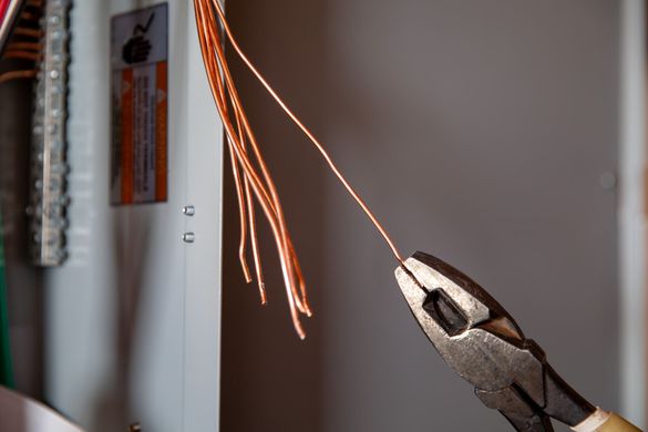 Pliers holding a copper wire in an electrical panel. Other wires are exposed nearby.