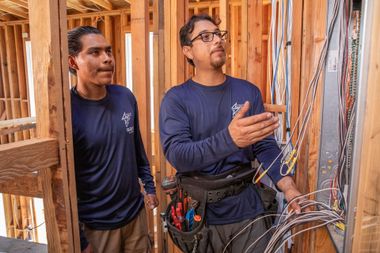 Two electricians inspecting wiring in a wooden construction frame. One points, the other looks on.