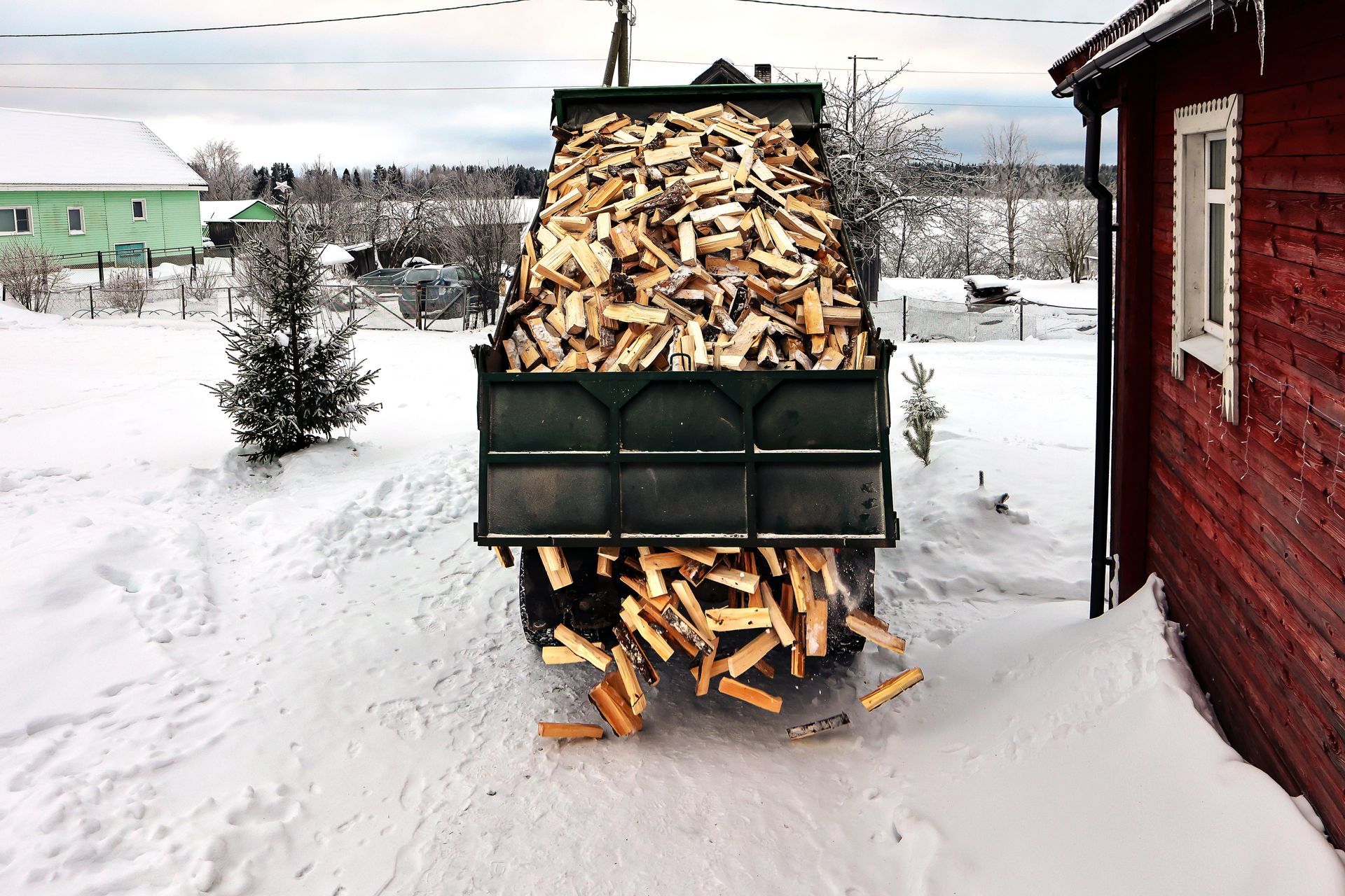 Pickup truck bed piled high with chopped firewood in a snowy yard beside a red building