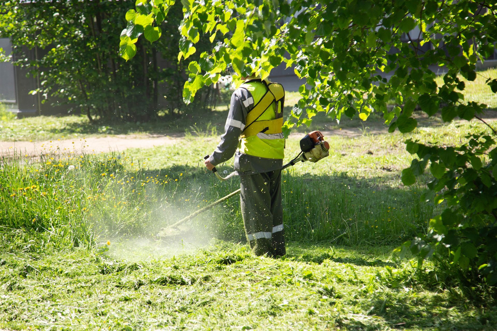 Person in a yellow vest using a backpack leaf blower to clear grass in a sunny park