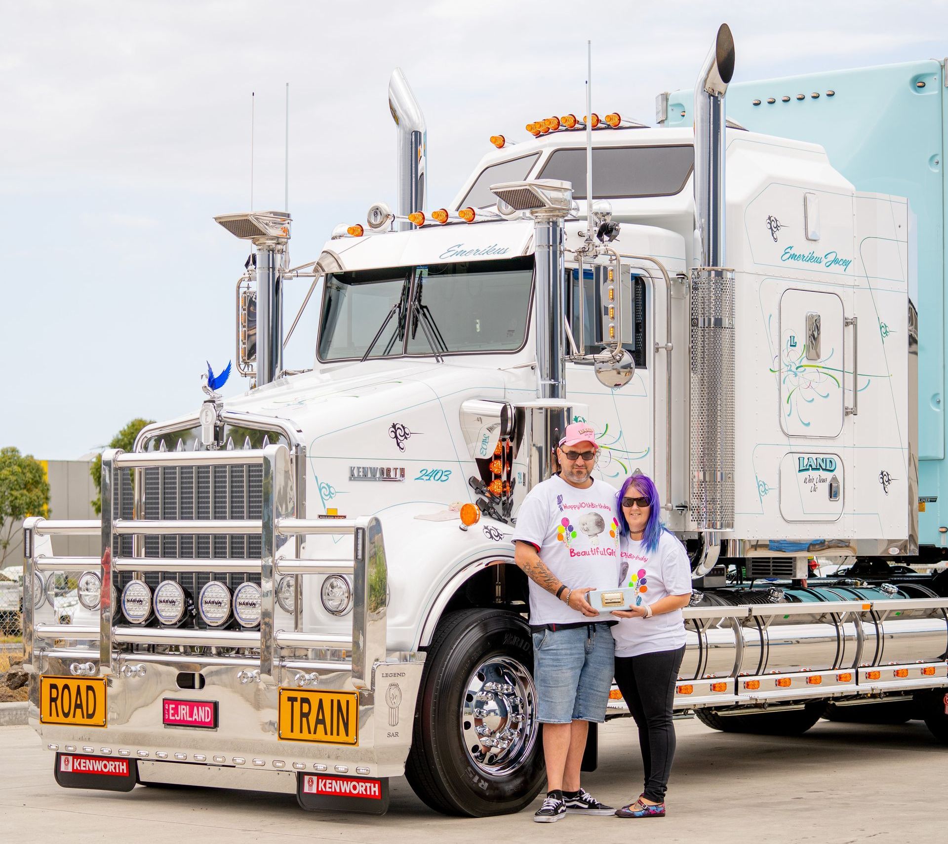 a man and a woman are standing in front of a semi truck that is decorated to support the Emerikus foundation.