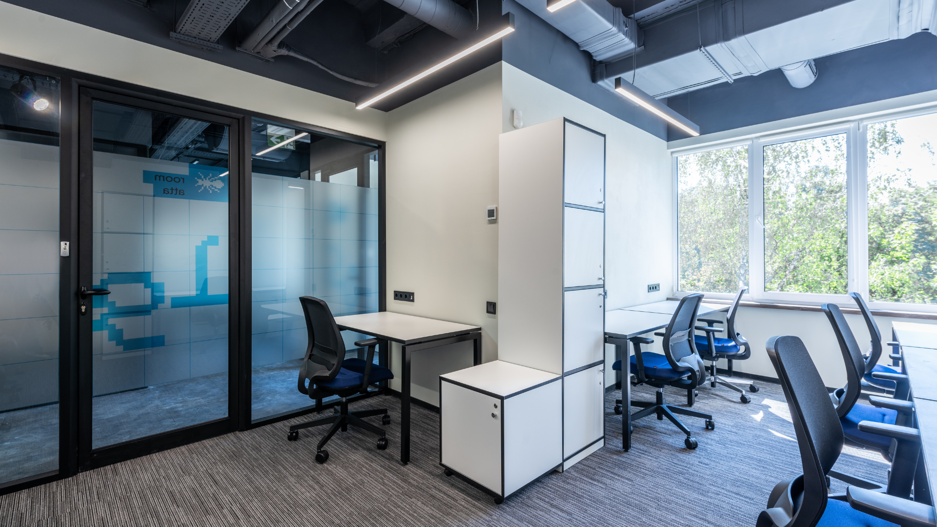 Modern office with glass-walled meeting room, desks, blue chairs, and large windows overlooking trees