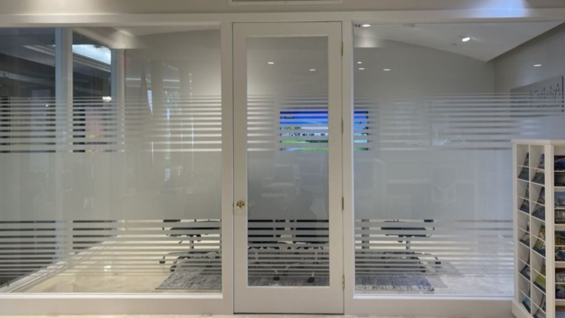 A white-framed glass office door and partition walls with horizontal frosted stripes, showing a meeting area inside.