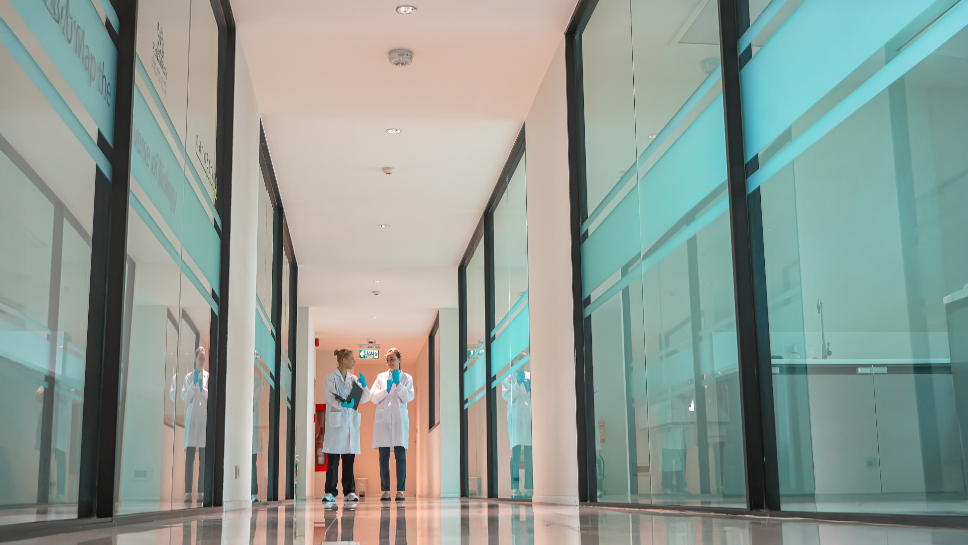 Two people walking in a bright glass-walled hallway with reflections on the polished floor.