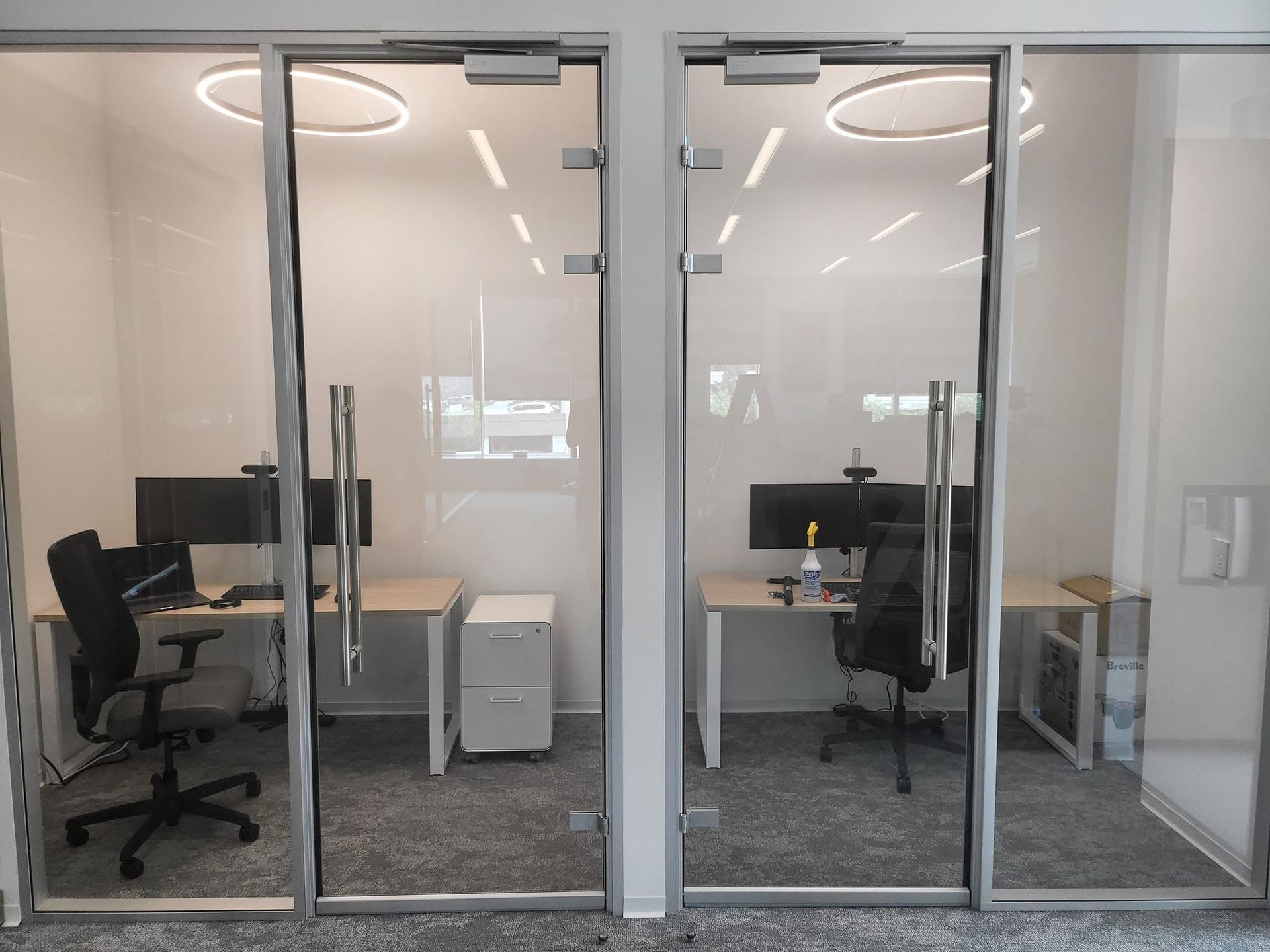Glass-walled office with desks, black chairs, computers, and a white cabinet inside