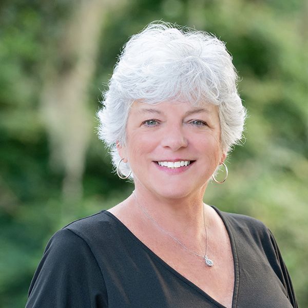 Smiling woman with short white hair in a black top, standing outdoors with a blurred green background