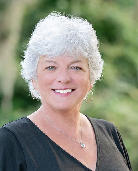 Whitney Hills with short white hair in a black top, standing outdoors with a blurred green background