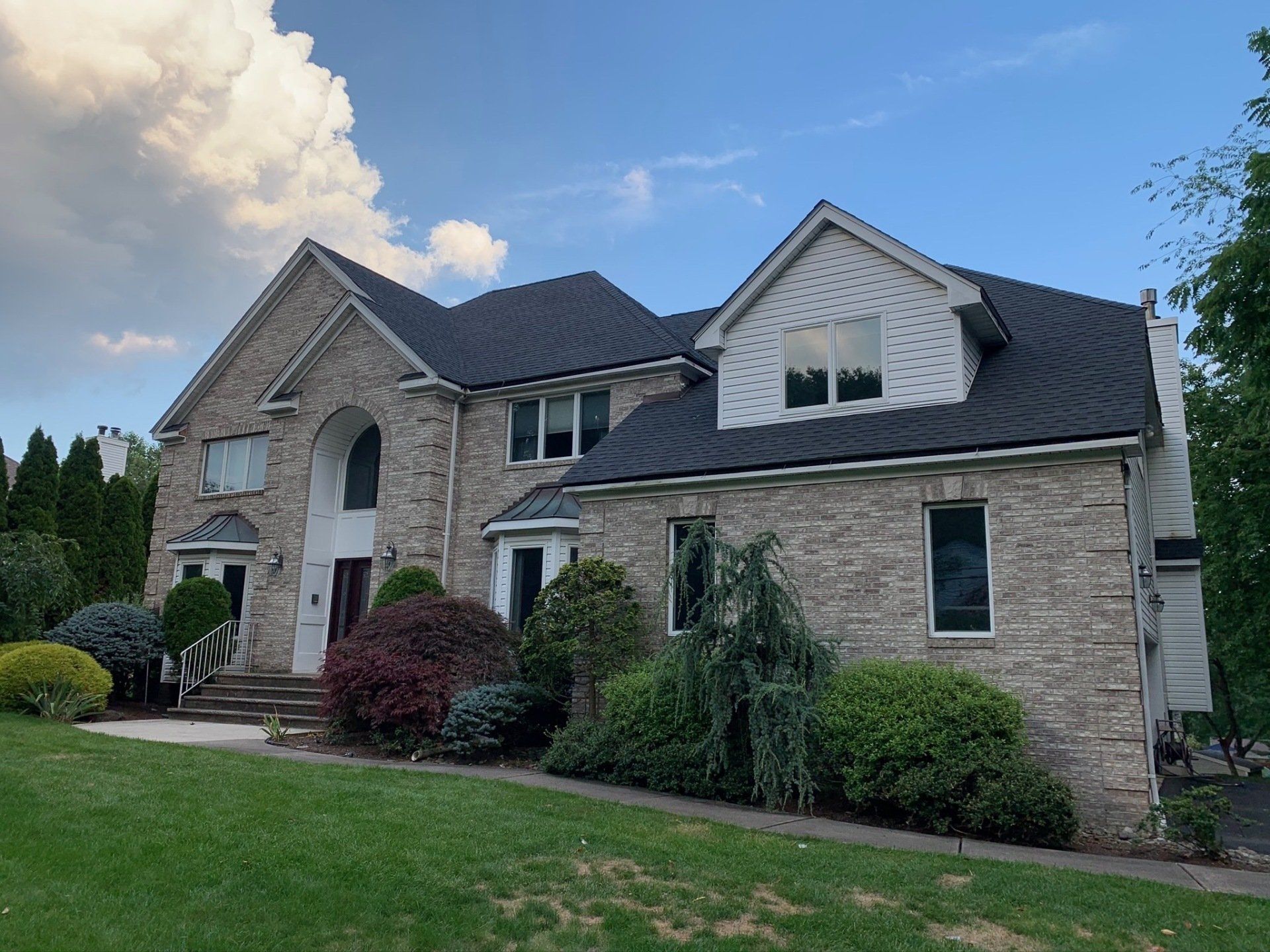 Large stone suburban house with dark roof, arched entry, and landscaped front lawn under a blue sky