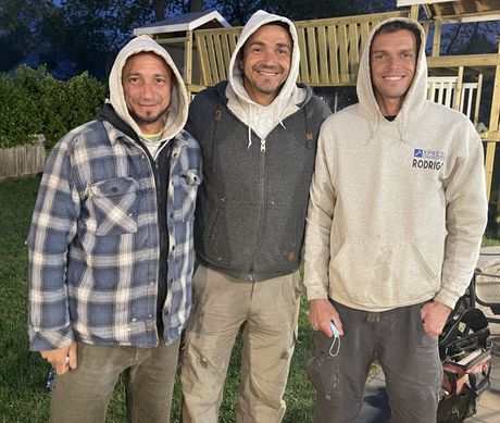 Three people in hoodies stand outdoors at night in front of a fence, smiling at the camera.