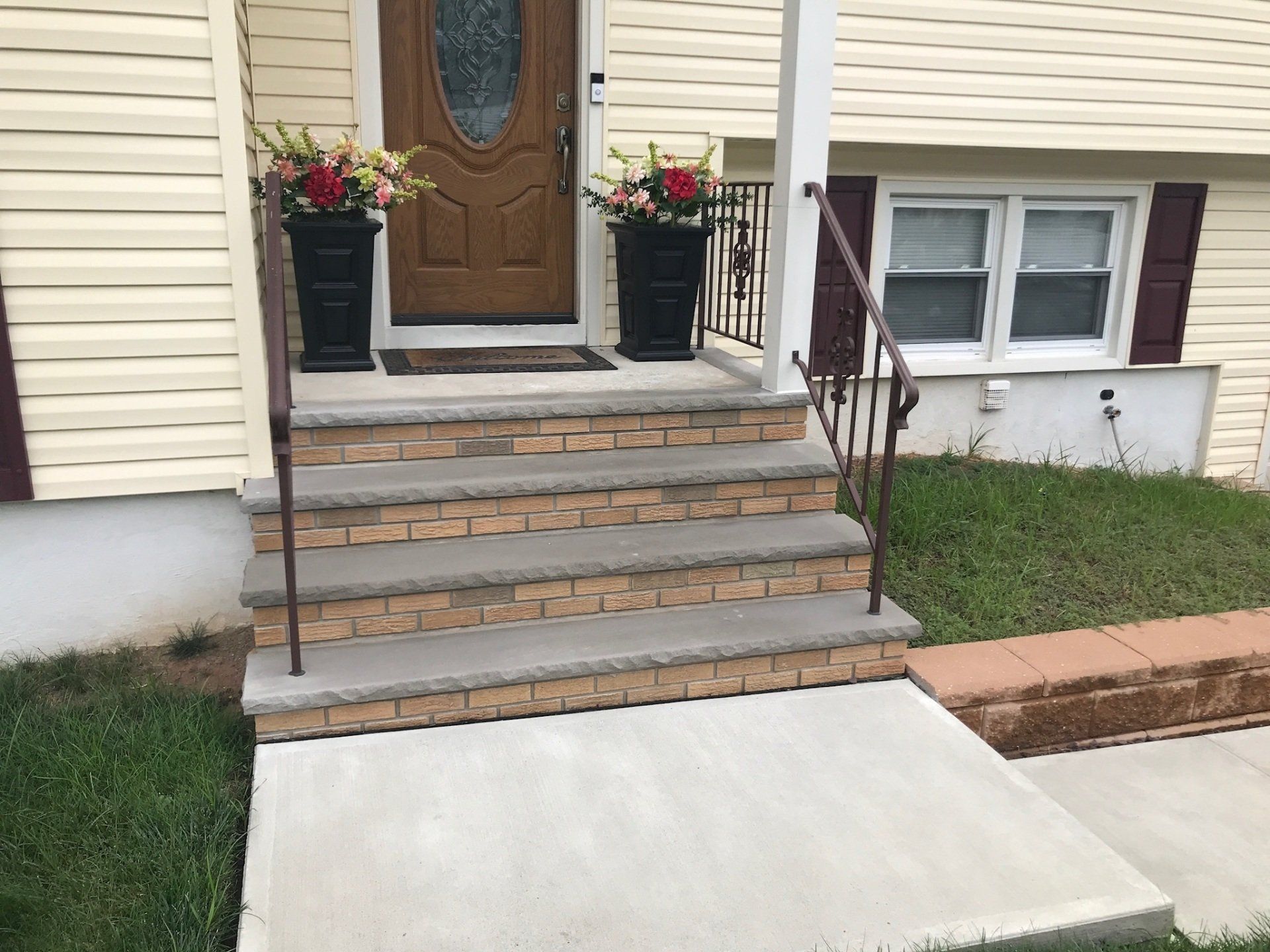 Front porch with steps, brown door, flower planters, and a concrete accessibility ramp.