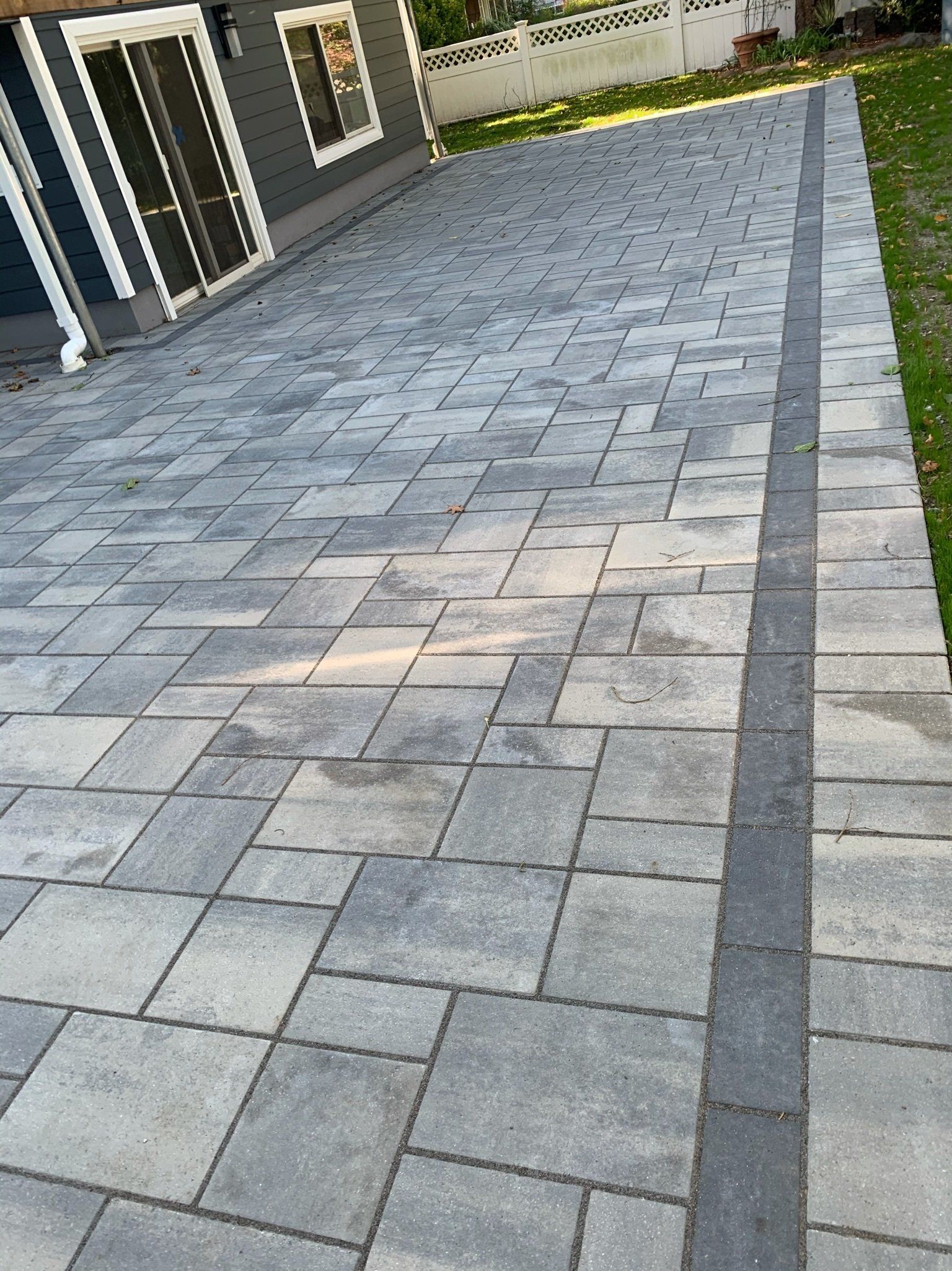 Gray stone-paved driveway beside a house, bordered by a strip of grass and a darker edge path.