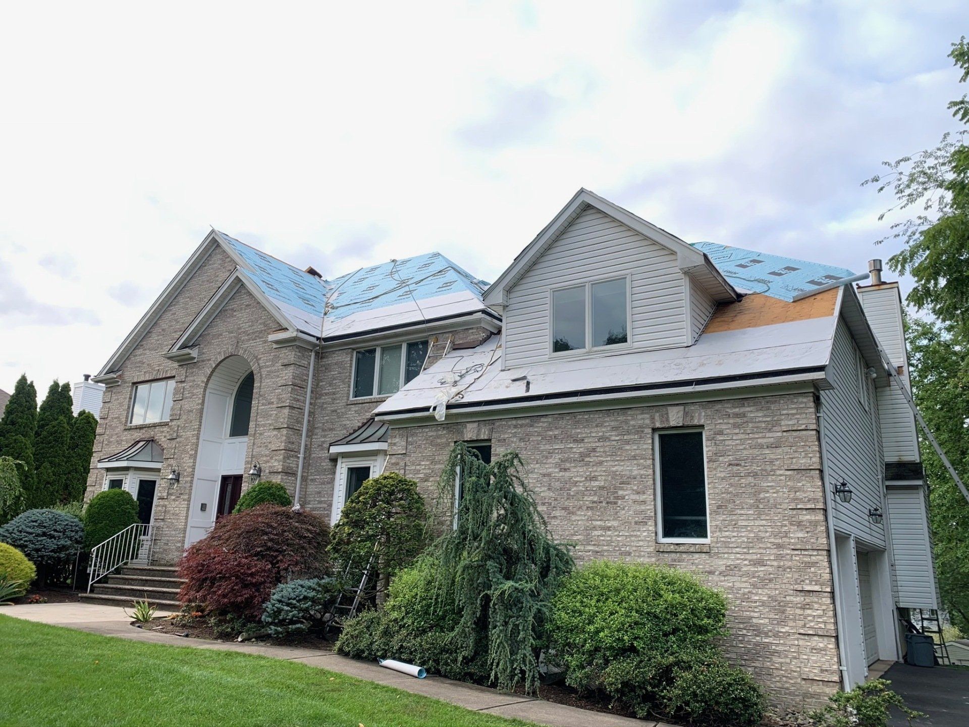 Large stone suburban house with a blue roof and front landscaping under a cloudy sky
