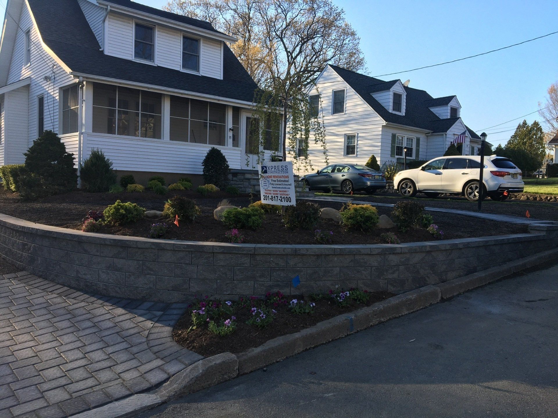 Residential street with white houses, landscaped stone wall, and parked white car on a sunny day
