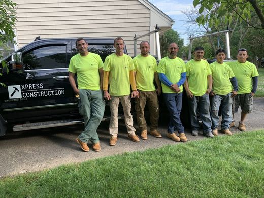 Seven workers in neon shirts standing by a black dump trailer in a driveway by a house