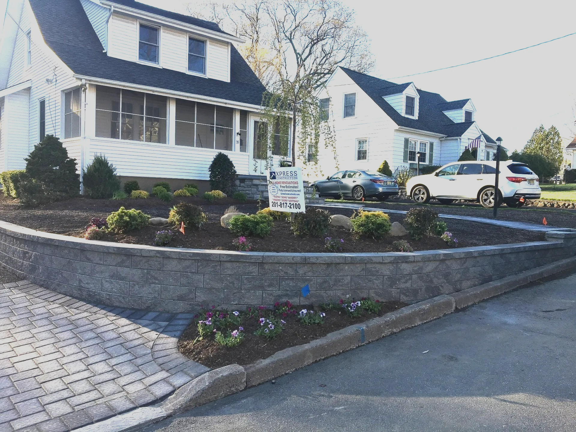 Residential neighborhood with white houses, a curved stone retaining wall, and a parked car on a paved street.