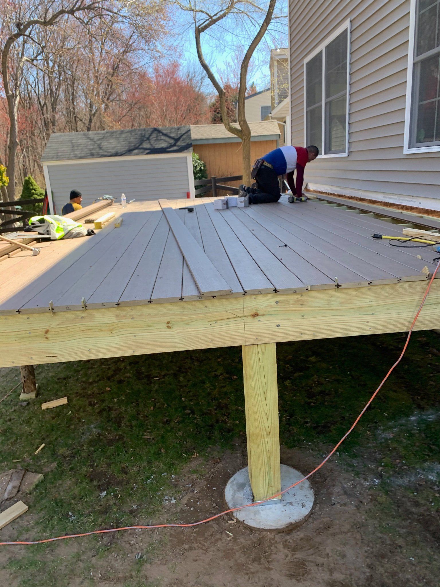 Workers installing gray deck boards on a raised wooden porch beside a house